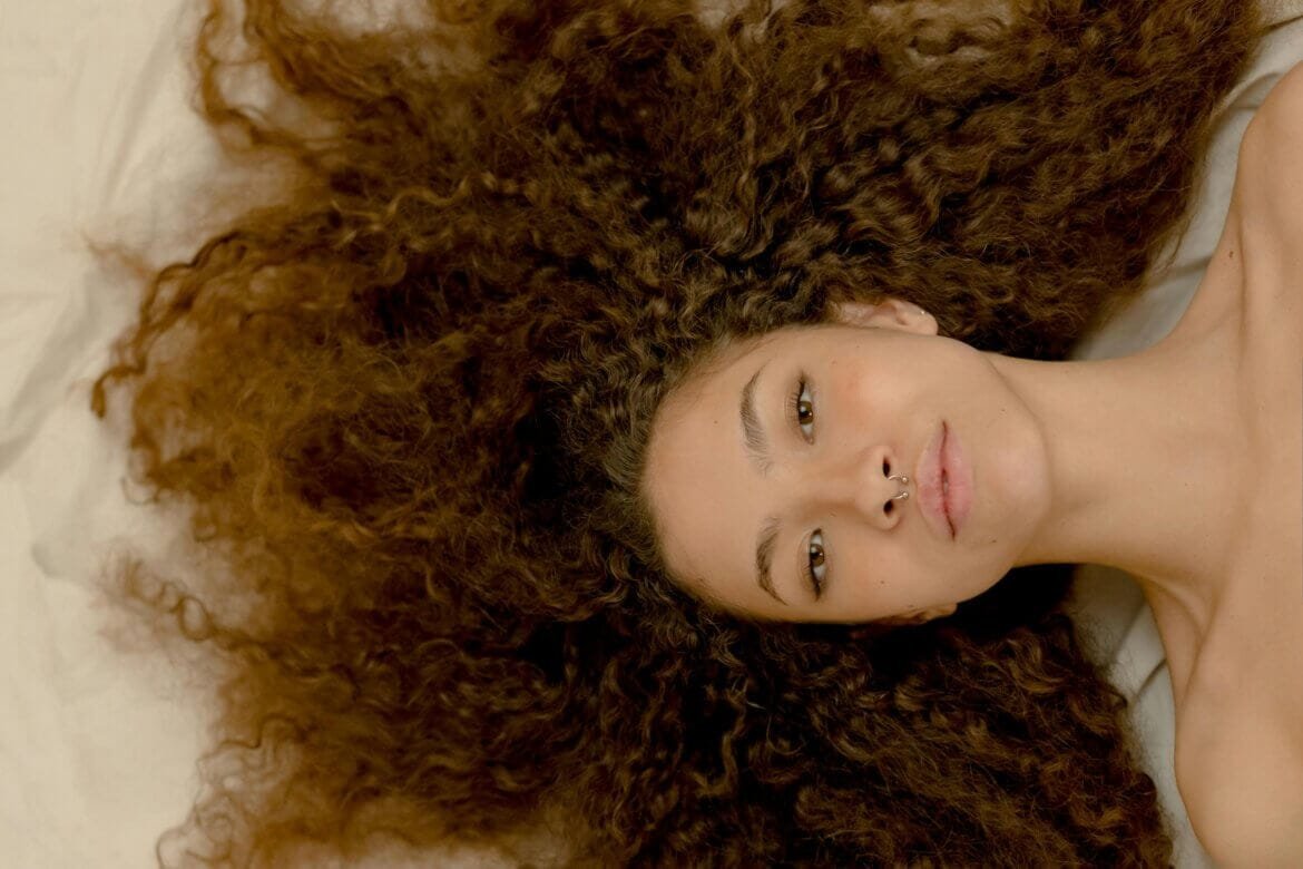A woman with curly hair lying down, looking directly at the camera.