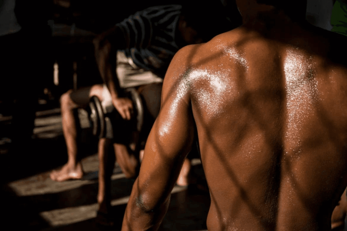 a muscular individual with a glistening, sweat-covered back in a gym-like setting, emphasizing physical effort and strength.