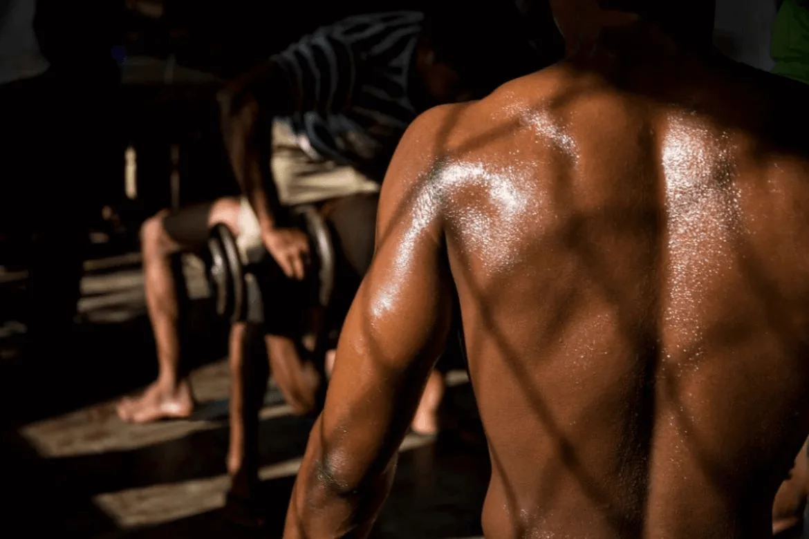 a muscular individual with a glistening, sweat-covered back in a gym-like setting, emphasizing physical effort and strength.