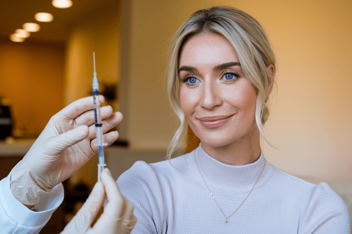 a blonde woman with blue eyes.In the foreground, a hand is seen holding a syringe.